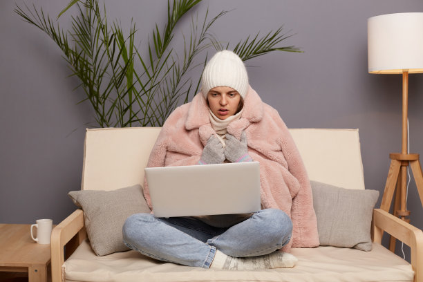 照片震惊惊讶惊讶寒冷的女人戴着帽子和冬天的外套坐在家里的沙发上，在笔记本电脑上工作，用大眼睛看着显示器。图片下载