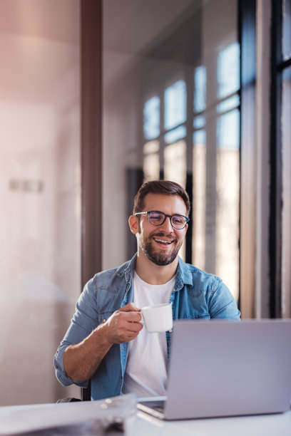 英俊的微笑的年轻人坐在办公室的窗户，喝咖啡，在笔记本电脑上工作。图片下载