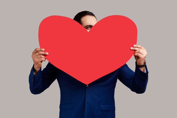 男人把脸藏在大大的红心后面，用好奇的眼神看着镜头，表达爱意。图片下载