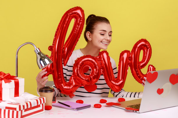 女人拿着爱的词箔气球，表达浪漫的情绪，看着笔记本电脑的屏幕。图片下载