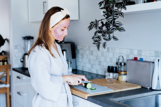 一名年轻的微笑女子用粉红粘土面膜切黄瓜片，制作清爽的眼膜。自然美容的程序，在家里皮肤护理。美容保健。有选择性的重点图片下载