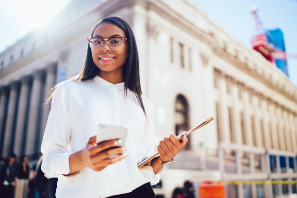 自信的黑人女性，戴着眼镜，拿着智能手机平板电脑和文件，走在市中心阳光明媚的街道上图片下载