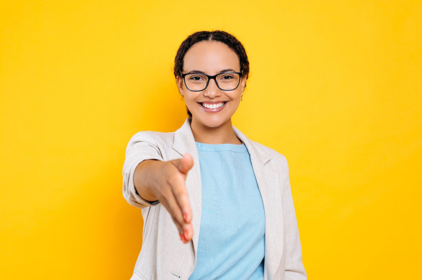 握手的姿势。积极愉快友好的巴西或拉丁美洲女性，商业伙伴，伸出手向前，做握手的手势，看着相机，微笑，站在孤立的黄色背景上图片下载