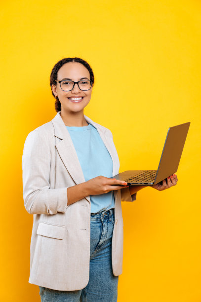 垂直照片的一个自信的积极漂亮的年轻卷发的女人，员工，在时尚的穿着，拿着一个打开的笔记本电脑，看着相机与愉快的微笑，站在孤立的黄色背景图片下载