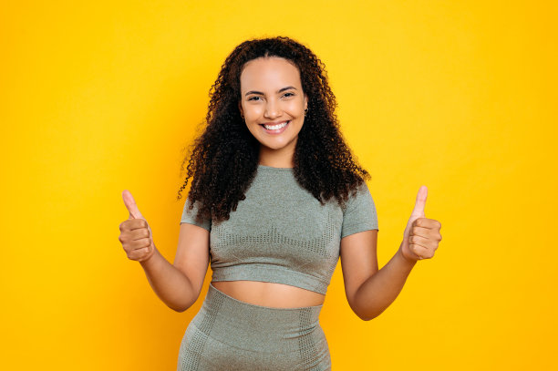 照片积极快乐可爱的卷发巴西或西班牙运动妇女在运动服，显示大拇指手势，看着相机，微笑，站在孤立的橙色背景图片下载