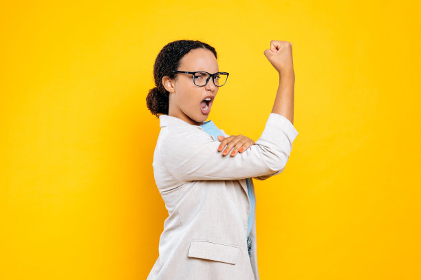 惊讶、兴奋、自信、强壮的年轻棕发西班牙或巴西女性，商务女士，展示二头肌，展示自己的力量，看着镜头，站在孤立的橙色背景上图片下载