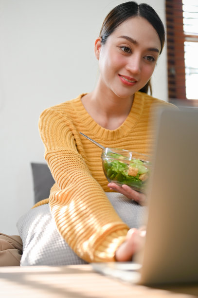 亚洲妇女拿着沙拉碗，吃沙拉蔬菜混合早餐，同时使用笔记本电脑图片下载
