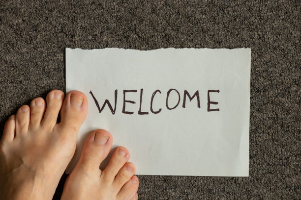一张写着欢迎语的纸放在灰色的地毯上，旁边是女人的腿，欢迎回家图片下载