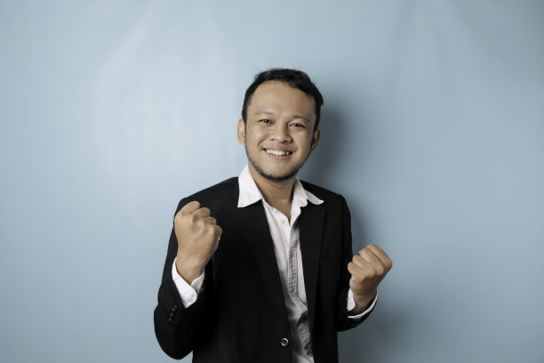 一名年轻的亚洲男子穿着被蓝色背景隔离的西装，表情快乐成功图片下载