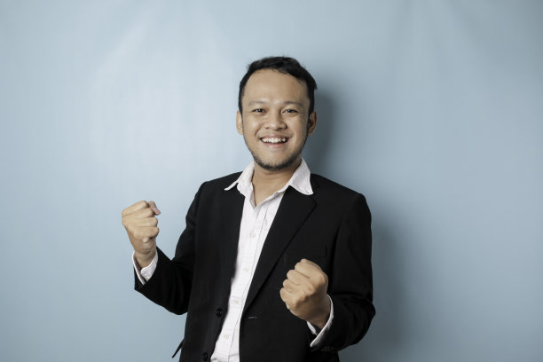 一名年轻的亚洲男子穿着被蓝色背景隔离的西装，表情快乐成功图片下载