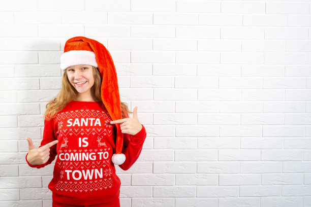 年轻微笑的金发女人在红色毛衣和圣诞老人的帽子指着手指，指着毛衣上的铭文图片下载