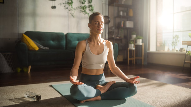 运动的年轻女性锻炼，练习冥想在早晨在她明亮的阳光房间在家里。穿着运动服的美丽女子在瑜伽垫上练习正念。图片下载