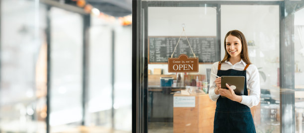 年轻友好的女性漂亮的白人咖啡店女老板围着围裙，开着牌子，小企业主微笑着，服务从平板电脑上的外卖应用程序带走订单。图片下载