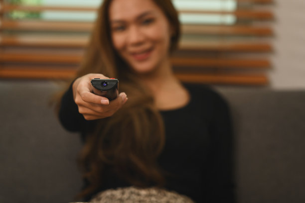 选择专注于手。年轻女子拿着遥控器看电视，在家里的沙发上放松。图片下载