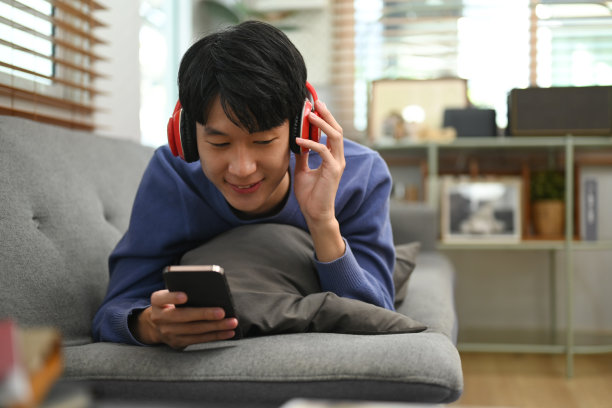 无忧无虑的年轻亚洲男子听音乐耳机和使用智能手机在家里的沙发上。图片下载