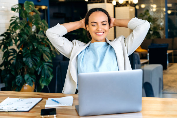 满意放松的混血女员工在工作时间休息，放松地坐在她的工作场所，双手放在脑后，对完成的项目感到满意，看着笔记本电脑，微笑友好图片下载
