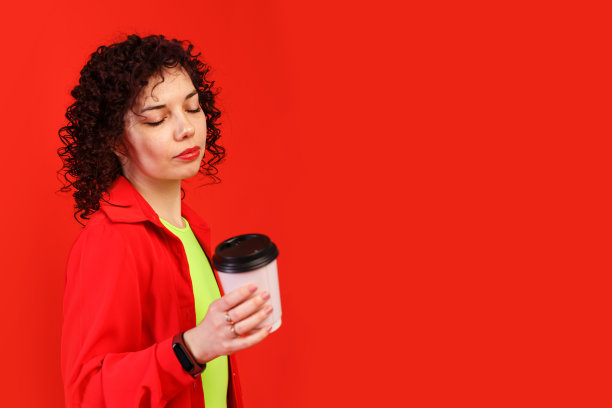 浪漫的年轻女子拿着粉色纸杯，盛着咖啡或茶。穿着亮红色衬衫和黄色t恤的卷发女孩。孤立在红色背景上。图片下载