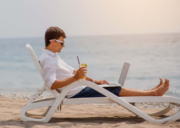 戴着墨镜，穿着白衬衫的年轻人正躺在海边的沙滩椅上，手里拿着鸡尾酒和笔记本电脑。图片下载