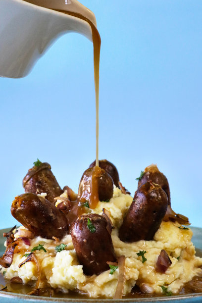 图片上的肉汁被从卤肉船倒在一盘儿童部分的传统英国香肠和土豆泥上，土豆泥和突出的猪肉香肠，上面覆盖着洋葱肉汁，丰盛的安慰食物，蓝色背景，重点在前景图片下载