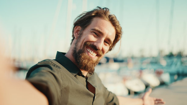 特写肖像的微笑男子与胡子聊天堤岸，在游艇的背景。正面特写的快乐年轻男性潮人使用手机图片下载