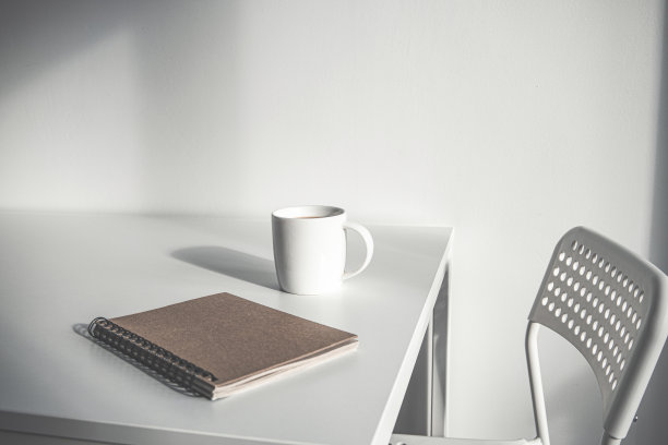 一杯白色的咖啡，白色的桌子上放着一个笔记本，旁边有一把椅子。图片下载