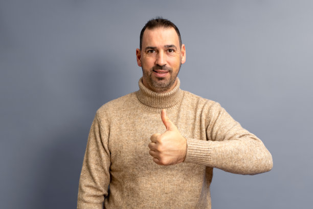 英俊的西班牙人穿着休闲毛衣灰色背景做快乐的大拇指手势与手。赞许的表情看着镜头显示成功图片下载
