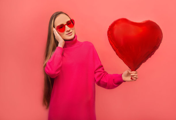 情人节和爱情概念:开朗的长发女孩，穿着粉红色连衣裙，戴着派对心形眼镜，手拿心形气球，看着相机孤立在五颜六色的粉红色背景上。图片下载