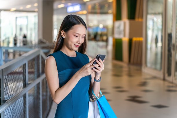 年轻的亚洲女商人女士，蓝色的裙子和购物袋使用她的手机在一个购物中心的室内自信图片下载