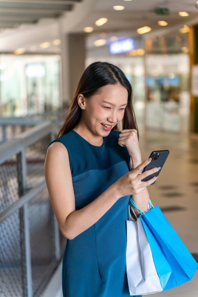 年轻的亚洲女商人女士，蓝色的裙子和购物袋使用她的手机在一个购物中心的室内自信图片下载