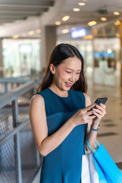 年轻的亚洲女商人女士，蓝色的裙子和购物袋使用她的手机在一个购物中心的室内自信图片下载