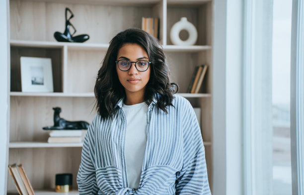 迷人的非裔美国女孩在休闲看着相机站在书架上的家。年轻的巴西女子戴着眼镜在酒店房间休息。成功的女企业家。业务。图片下载