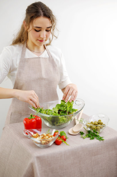 年轻的女孩用手混合生菜，她倒进玻璃盘子，素食，木勺，围裙，桌布，白色背景，年轻的女子少年准备晚餐，家庭晚餐，早餐，午餐图片下载