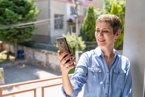 一个女人成熟的白人女性，在明亮的一天或早上，在家里的窗户边使用手机，日常生活，快乐的微笑，快乐和自信，自拍照片或打视频电话复制空间图片下载