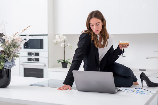 女商人坐在厨房桌子上，手里拿着玻璃杯和笔记本电脑图片下载