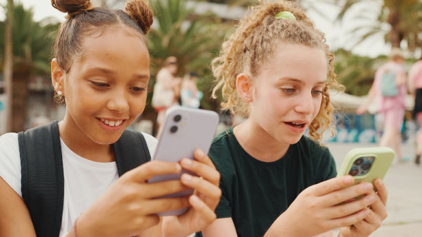 两个十几岁前的女孩朋友坐在海滨看视频，照片，在户外的城市景观背景下发布在社交网络上。图片下载
