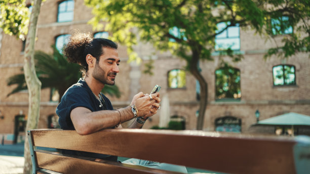 迷人的意大利男子与马尾和胡茬正在使用手机坐在长凳上在老建筑背景。型男写留言，用智能手机看照片图片下载