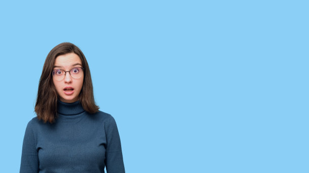 一个惊讶的学生女孩的肖像，在眼镜和一件毛衣孤立在蓝色背景上张开嘴图片下载