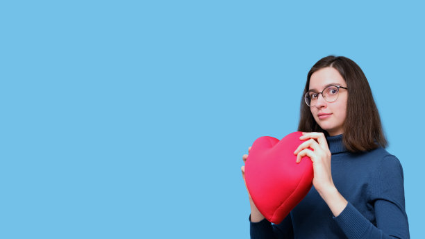 一个美丽的学生女孩的肖像，戴着眼镜，抱着一个明亮的红色心形枕头在蓝色的背景图片下载
