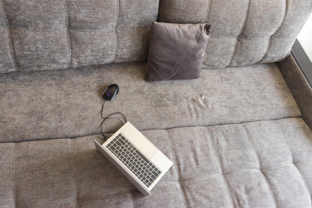 黑色的电脑鼠标和一台钢制薄笔记本电脑放在灰色的阁楼沙发上。远程工作和在家上网的概念。没有人。图片下载
