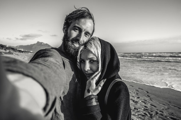 相爱的旅行情侣在日落时在荒芜的海滩上自拍-女人有磁性的凝视和她的时髦男友在寻找自拍相机时玩得很开心-黑白编辑图片下载
