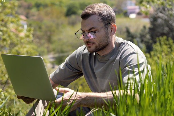 戴眼镜的男人坐在绿草地上用笔记本电脑工作。图片下载