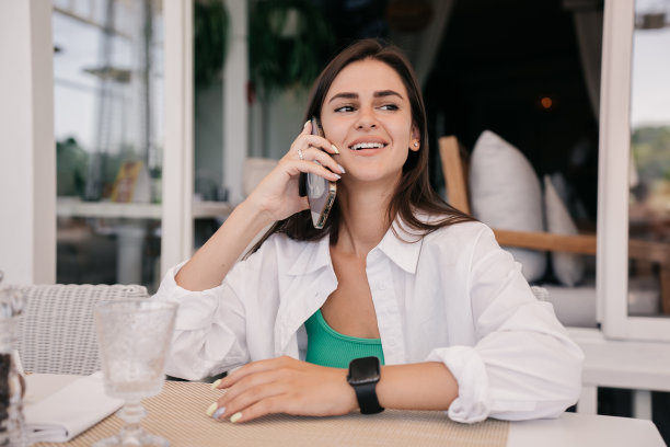 可爱的西班牙褐发年轻成年女子在白衬衫坐在桌子上的咖啡馆打电话与男朋友看一边。漂亮的白人女孩在约会前兴奋。夏季。情人节。图片下载