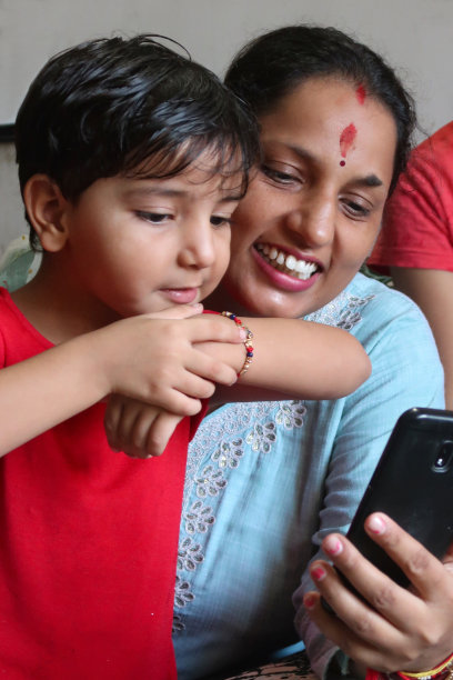 特写图片，印度女孩坐在她母亲的大腿上，视频中女人手里拿着智能手机打电话，女人在家庭房间里穿着Kameez(束腰外衣)传统服装，头部特写，重点在前景图片下载