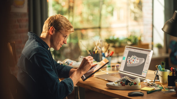 经验丰富的男性青少年设计师工作与数字图形平板电脑触摸屏，绘图草图的一个新的独特的鞋子设计。他的产品的3D原型在编辑软件上的笔记本电脑显示。图片下载