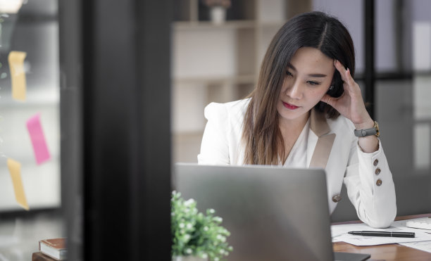 沮丧的年轻女子感到疲惫和压力，手摸她的头，而坐在她的工作场所。图片下载