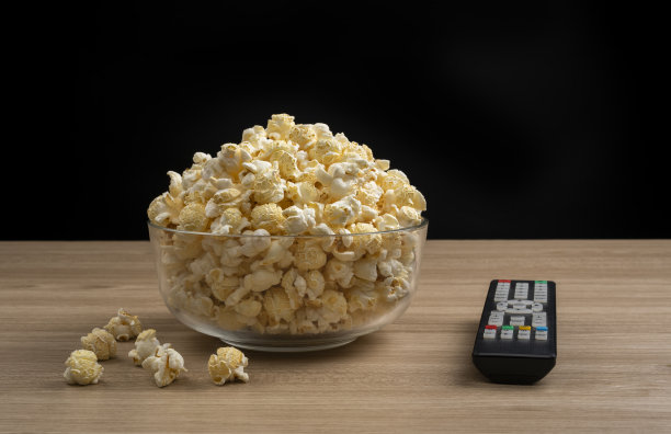 一个玻璃碗的爆米花和电视遥控器在木桌，黑色背景。图片下载