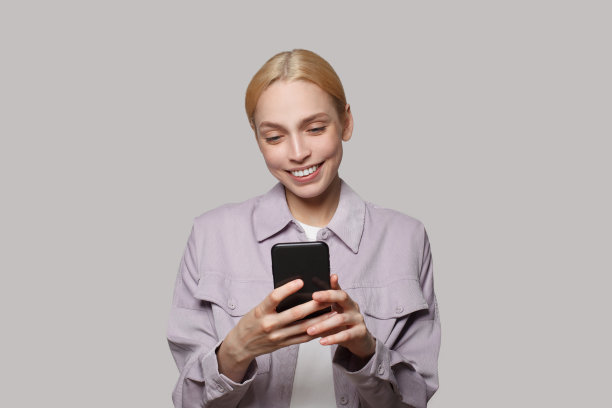 开朗的年轻女子与金发手持智能手机在白色的工作室墙壁背景。人们生活方式的概念。模特用手机，打短信图片下载