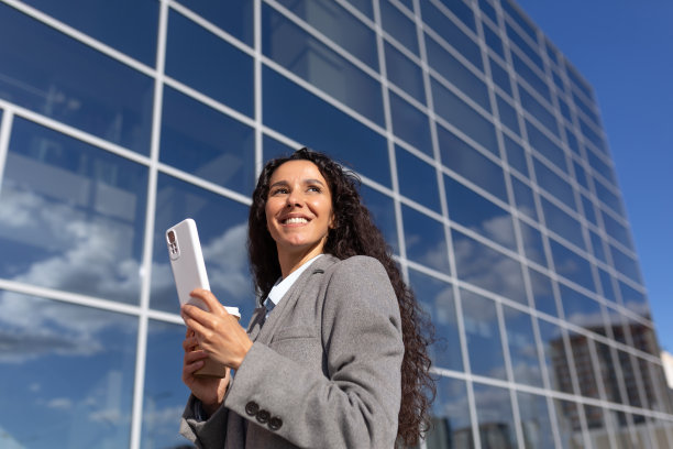 一位穿着西装的年轻拉丁美洲女商人站在办公室附近，拿着手机微笑图片下载