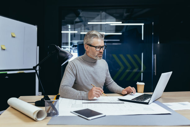 头发花白的资深建筑师坐在办公室里，用笔记本电脑工作，绘图。使笔记图片下载