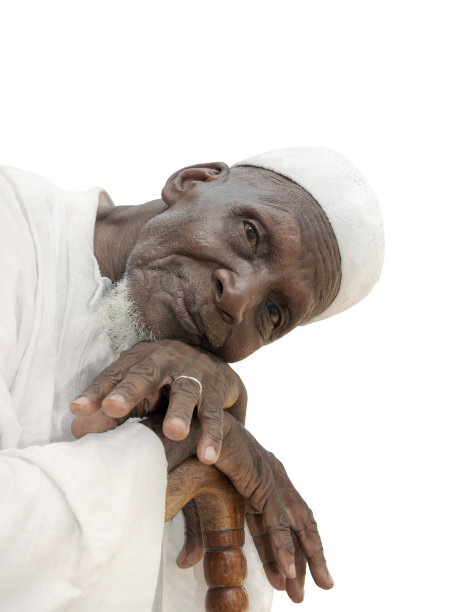 非洲老人坐着看镜头，表情嬉笑，庆典服装和帽子，八十岁高龄，孤立(剪掉)，白色背景，照片图片下载